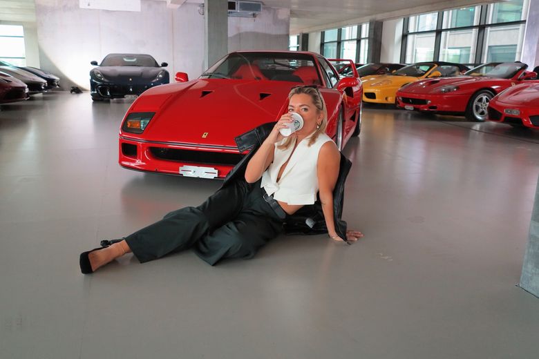 Danni Menzies sitting on the floor of a Ferrari showroom with a coffee, an F40 behind her.