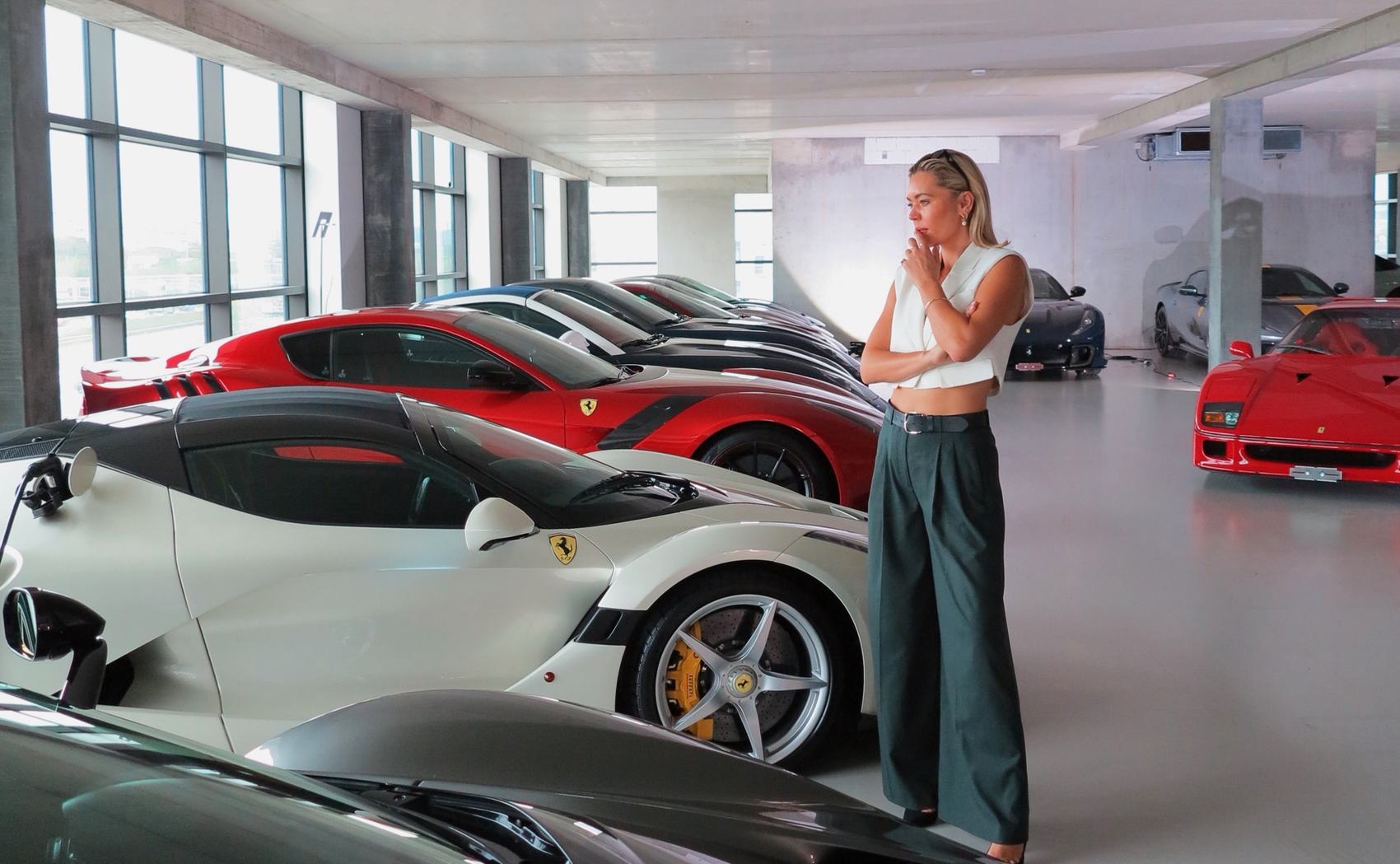 Danni Menzies standing among a collection of Ferraris in a concrete showroom.